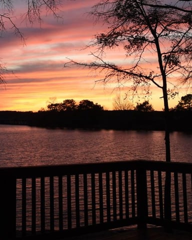 Sunset over a lake viewed from a wooden deck railing, with bare trees silhouetted against orange and pink sky