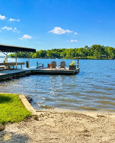 Adirondack chairs arranged around fire pit on sandy beach overlooking calm lake with forested shoreline