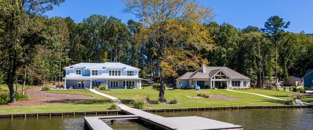 Lakeside pool and hot tub with lounge chairs, umbrella, and stone house overlooking blue water and forested shoreline
